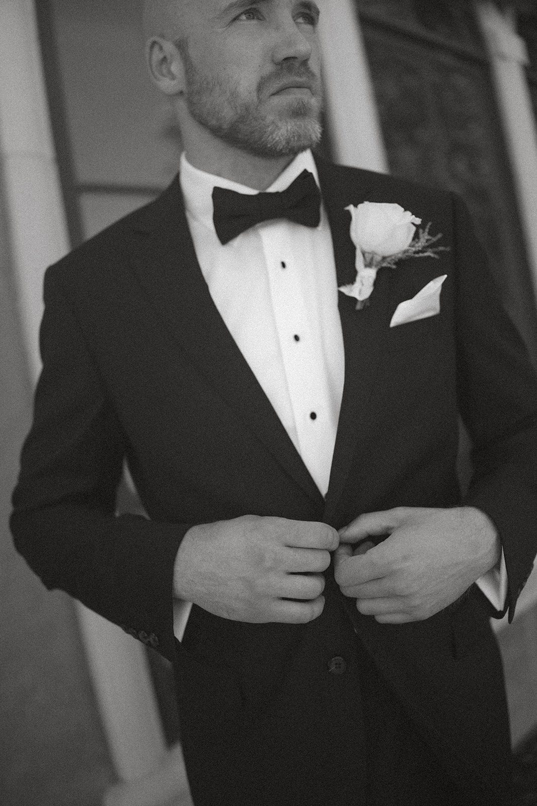 Black-and-white portrait of a groom adjusting his black tuxedo with a white rose boutonniere before his san luis obispo wedding.