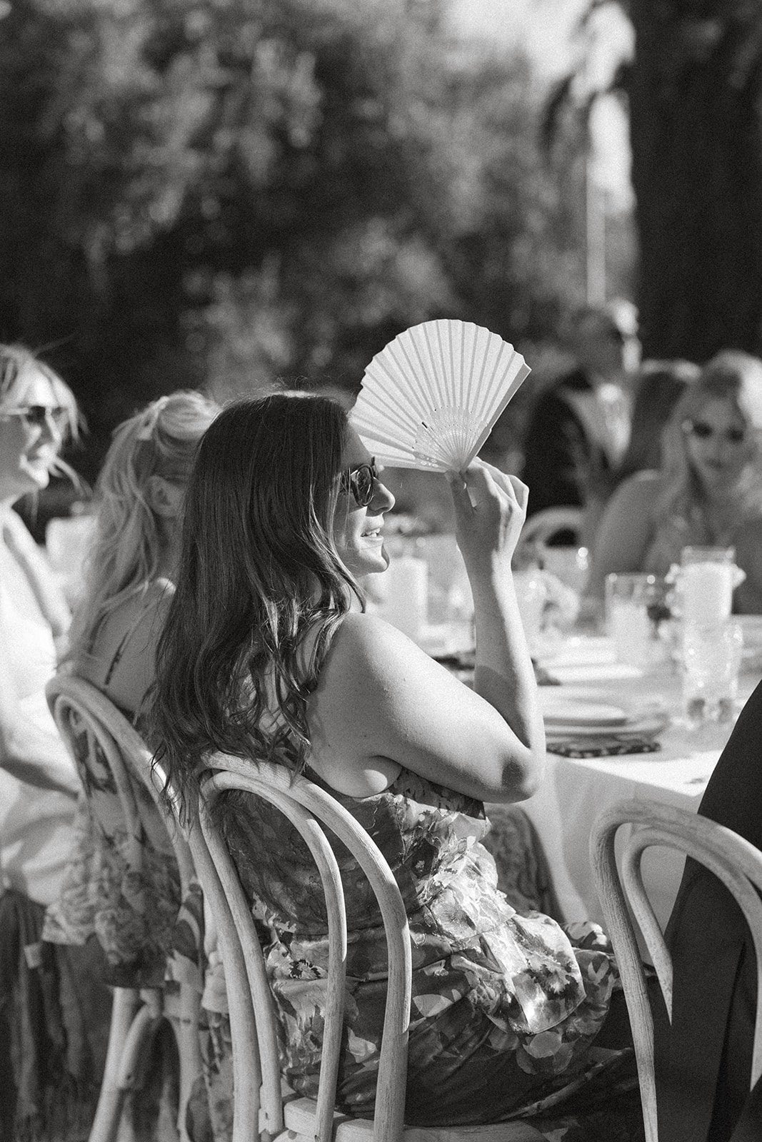 Black-and-white candid of a wedding guest in sunglasses holding a white folding fan during an outdoor vineyard wedding reception.