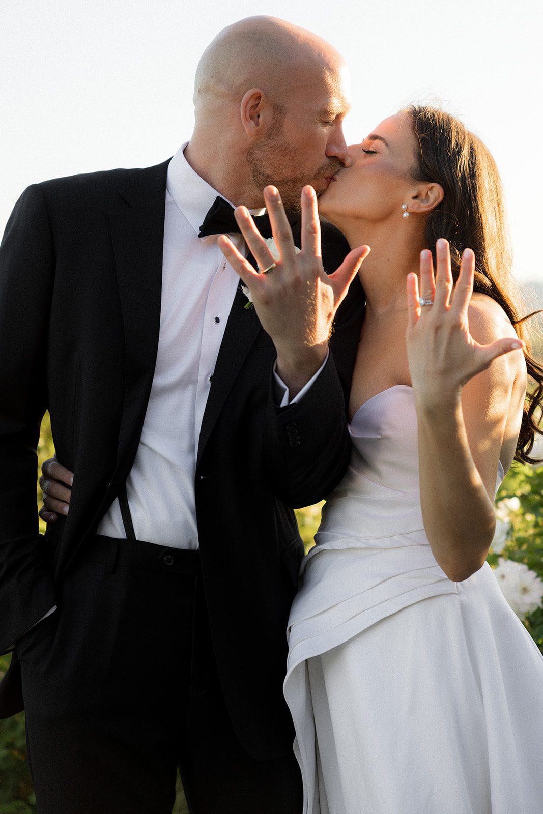 Bride and groom kiss while holding up their hands to display their wedding rings during a golden-hour portrait session at Villa San Juliette.