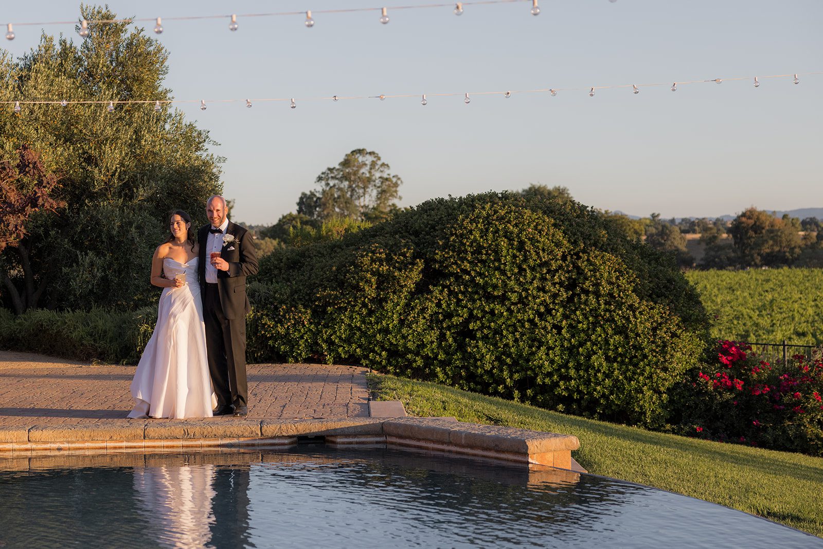 Bride and groom stand together holding drinks beside a reflecting pool beneath string lights, surrounded by vineyard greenery at Villa San Juliette.