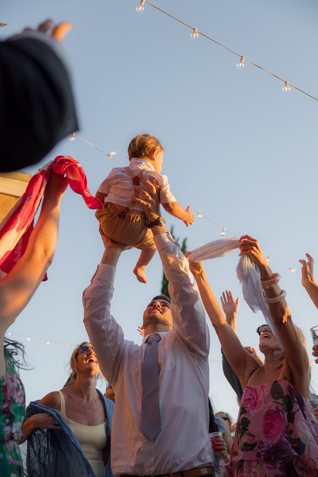 Wedding guest lifting a toddler into the air as friends wave colorful scarves beneath string lights at a Villa San Juliette wedding.