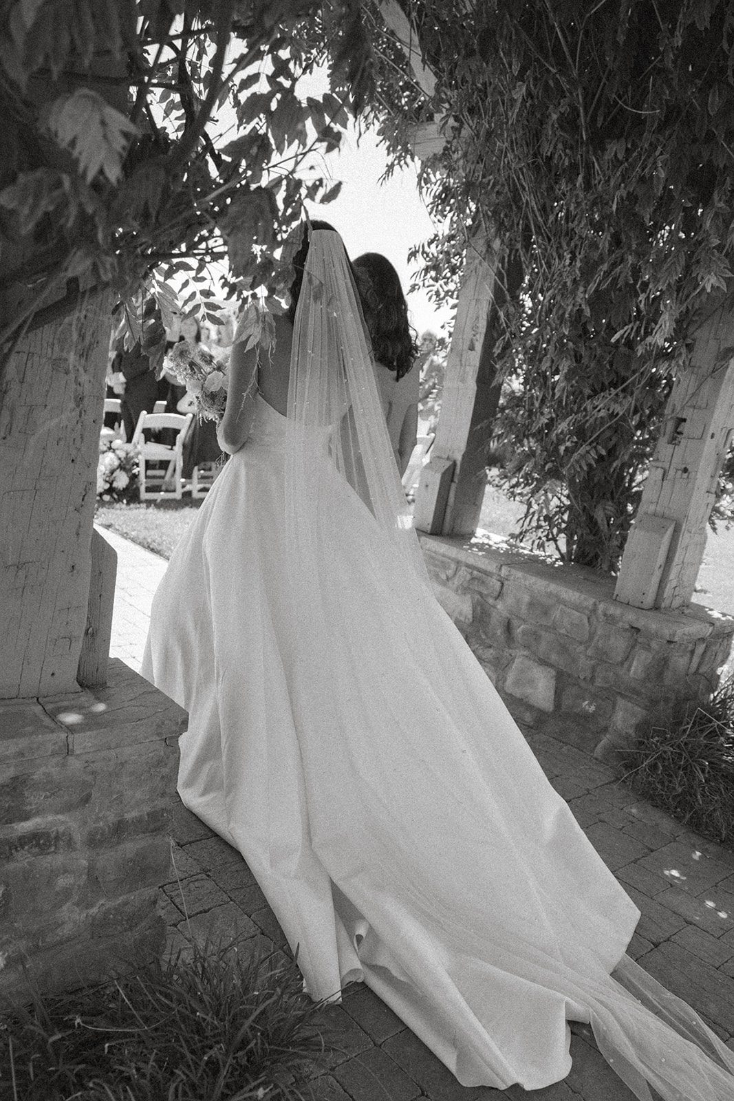Black-and-white photograph of a bride in a flowing satin wedding gown and long veil walking toward her outdoor Villa San Juliette wedding ceremony.