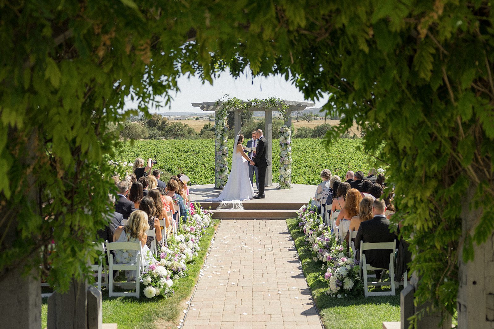 Bride and groom exchanging vows beneath a floral-covered pergola overlooking the vineyards at their Villa San Juliette wedding.