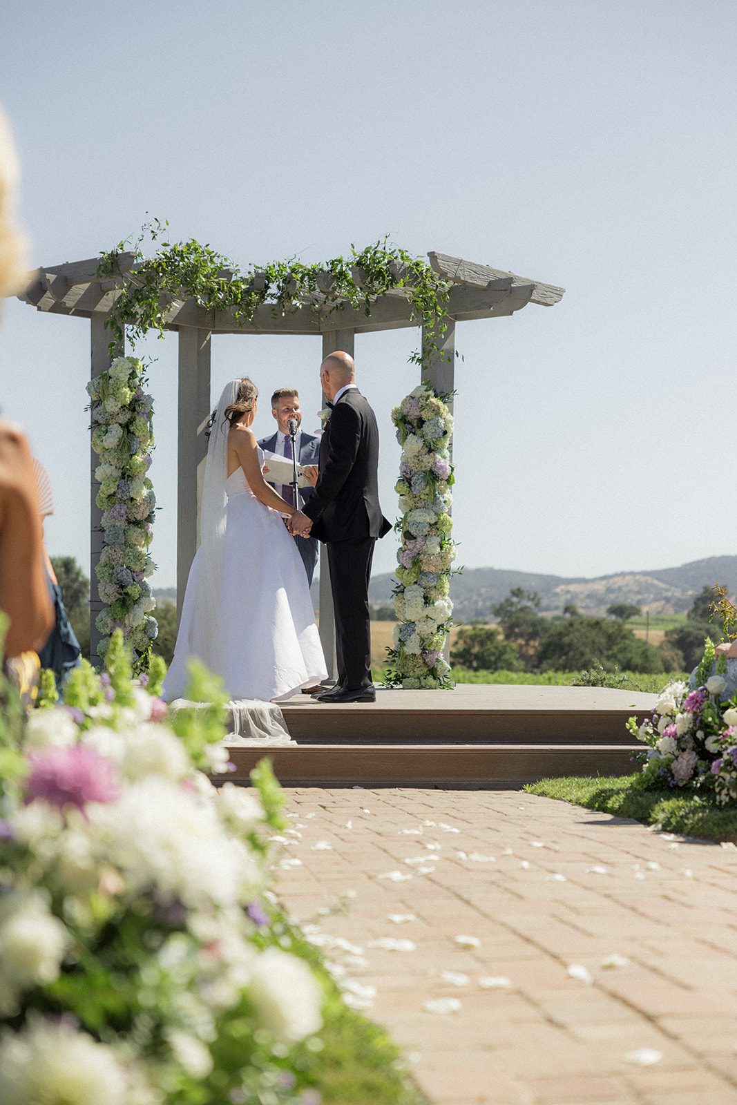 Bride and groom holding hands beneath a floral-covered pergola during their Villa San Juliette wedding ceremony overlooking rolling hills.