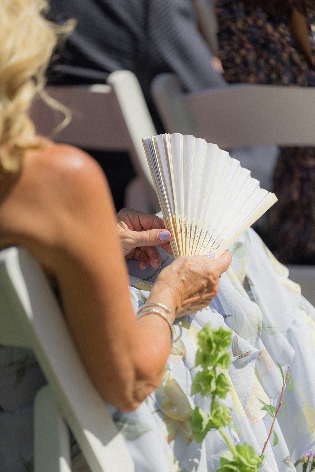 Close-up of a wedding guest holding a white folding fan during a sunny outdoor ceremony at a vineyard wedding venue.