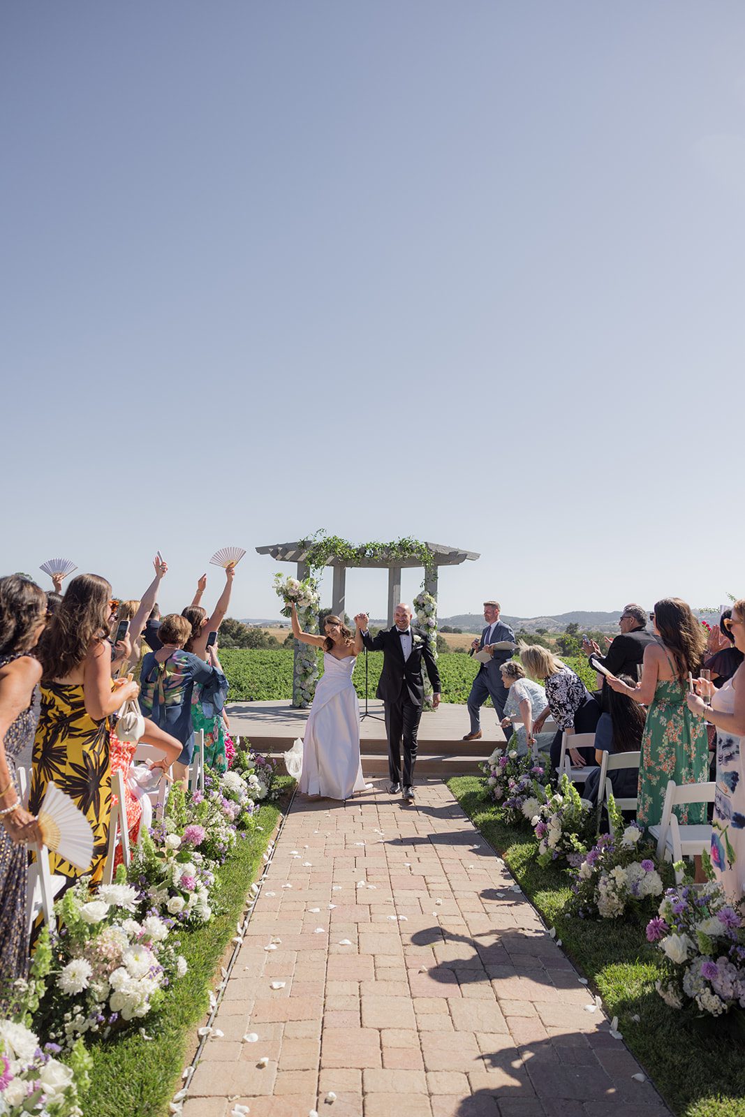 Newlyweds celebrating down a flower-lined ceremony aisle with vineyard views at their Villa San Juliette wedding.
