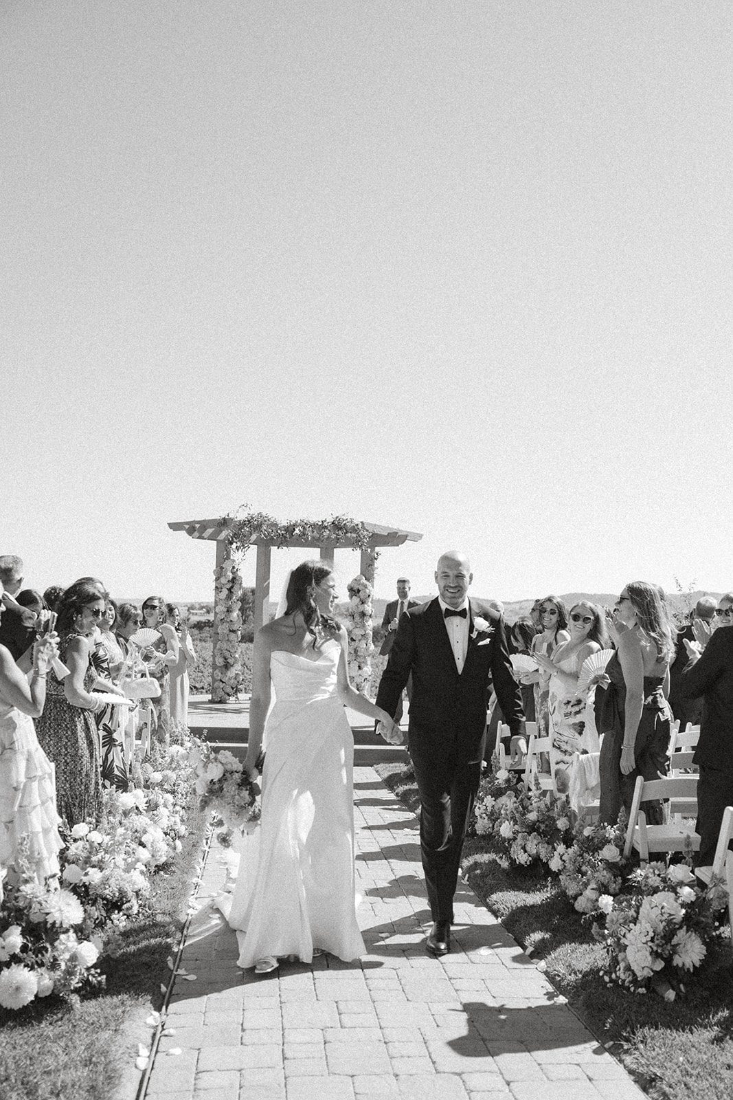 Black-and-white photograph of newlyweds walking hand in hand down a flower-lined aisle as guests applaud at their Villa San Juliette wedding.