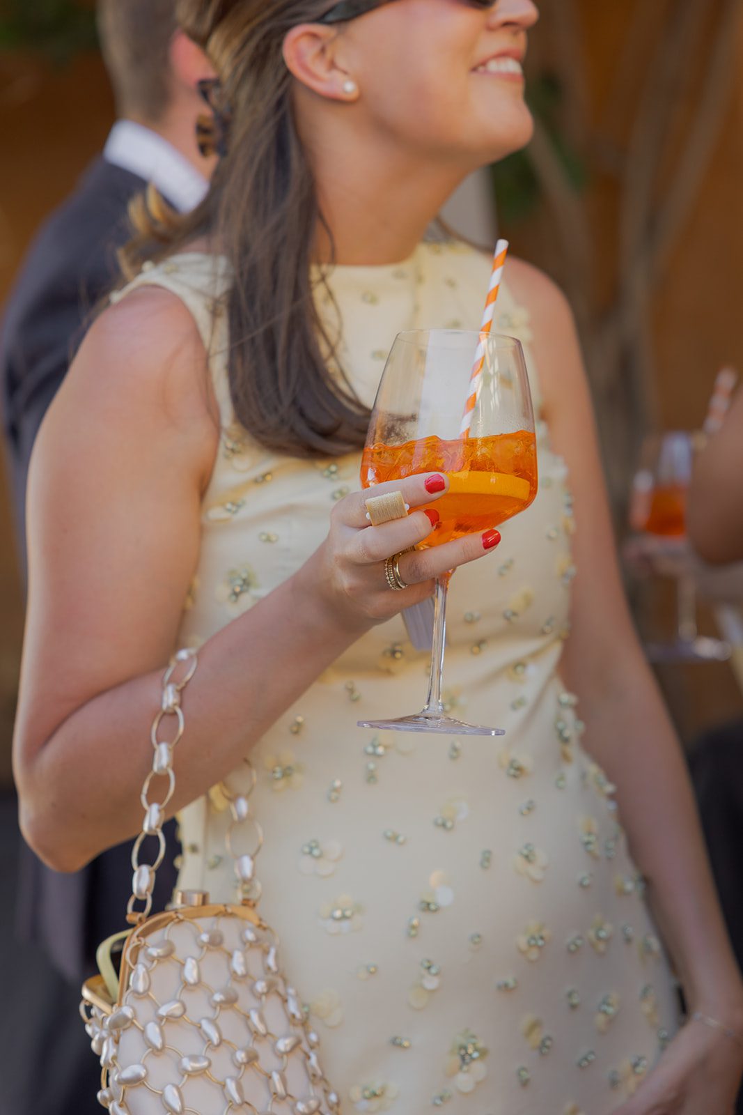 Wedding guest in a cream embellished dress holding an orange spritz cocktail with a striped straw at a Villa San Juliette wedding.