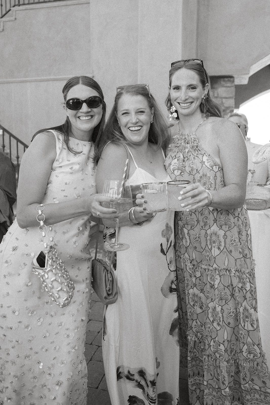 Black-and-white candid of three wedding guests in patterned dresses smiling and raising cocktails during a san luis obispo wedding celebration.