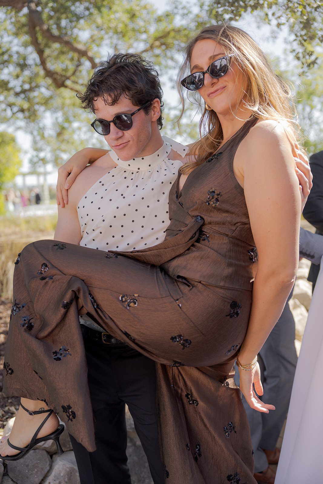 Two wedding guests wearing sunglasses pose together, with one playfully lifting the other in a brown floral dress at a Villa San Juliette wedding.